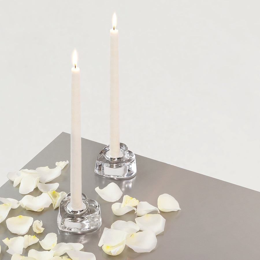 Two lit tealight candles sit in Orrefors: My Heart Votive Candle Holders on a gray surface, surrounded by scattered white rose petals.