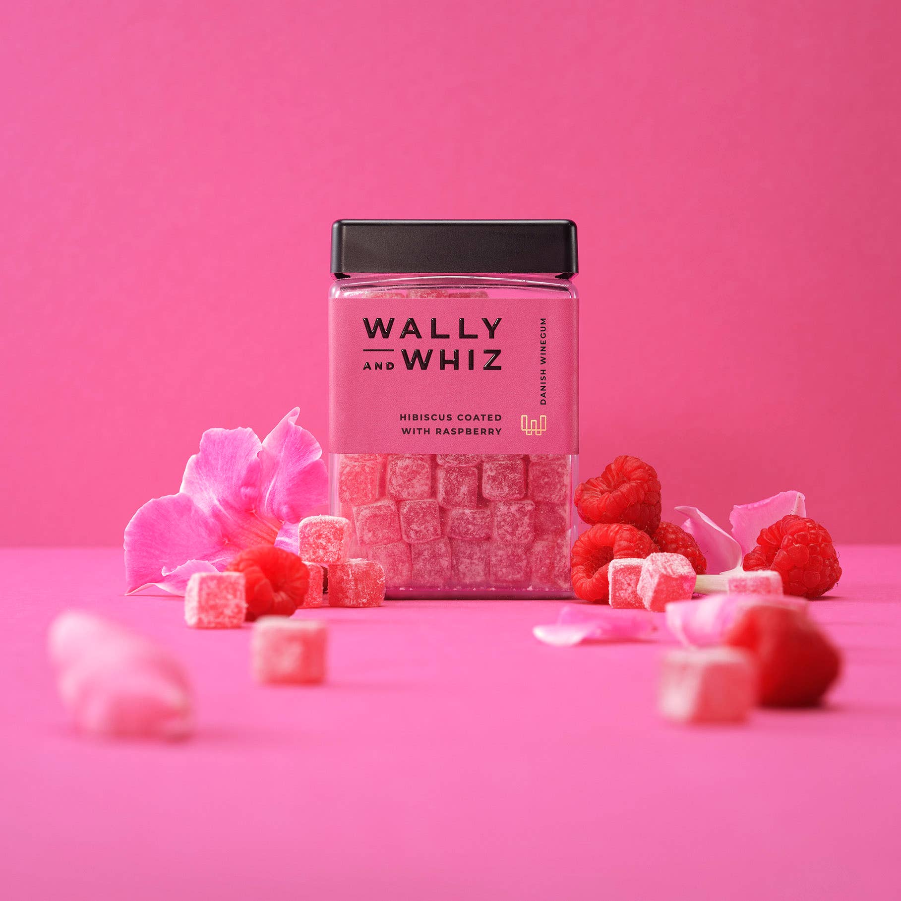 A jar of Wally & Whiz Danish Winegum Candy Gummies in hibiscus-coated raspberry flavor is displayed with pink flower petals and raspberries on a pink background.
