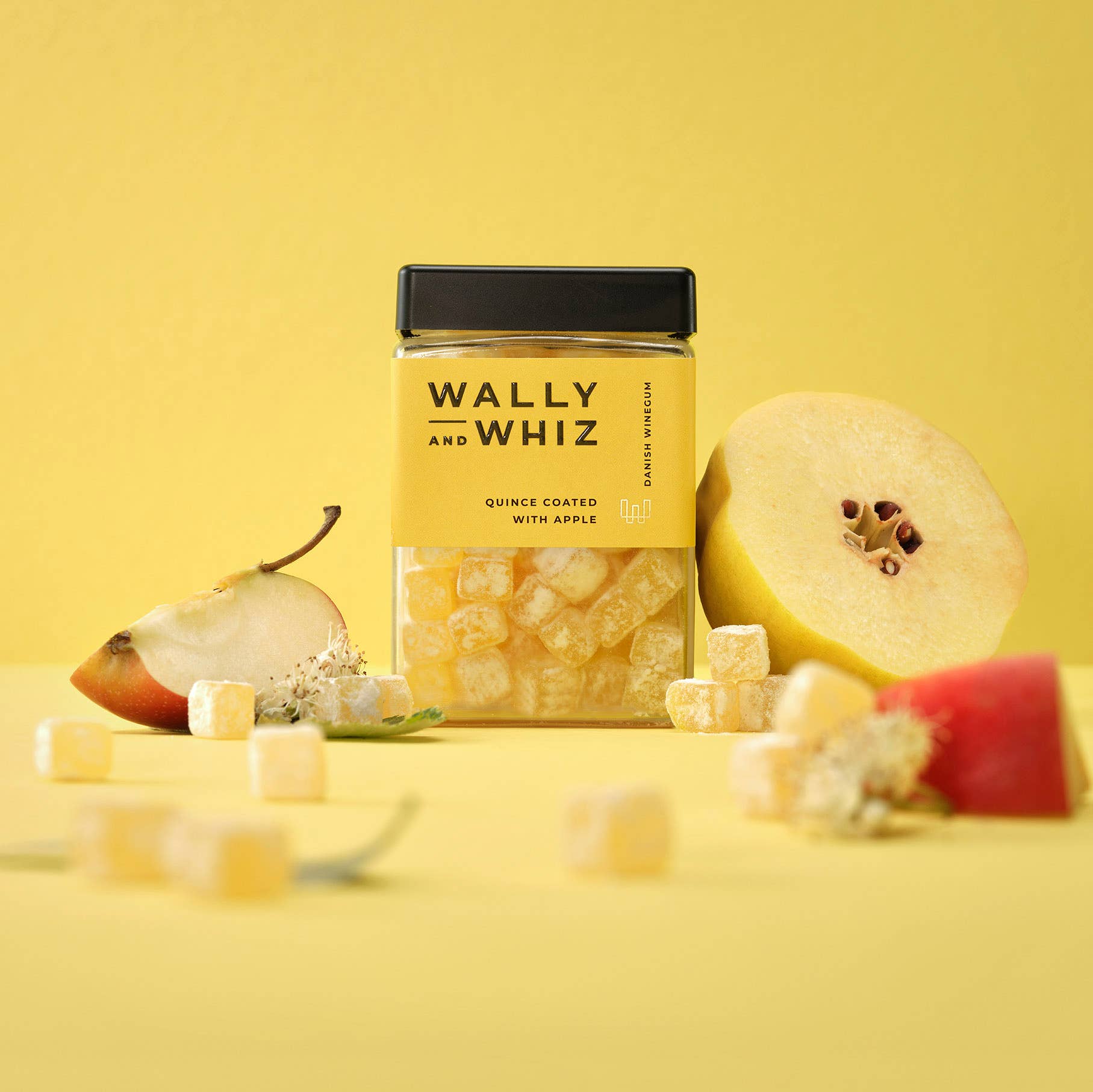A jar of Wally & Whiz - Danish Winegum Candy Gummies is displayed among fruit cubes, quince, and pear slices on a yellow background—delicious vegan and gluten-free gummies perfect for a tasty treat.