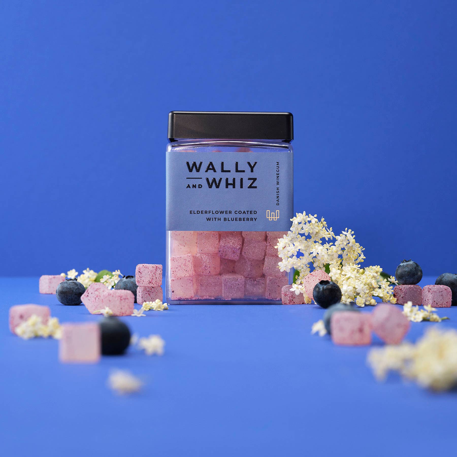 A clear jar labeled Wally & Whiz - Danish Winegum Candy Gummies contains pink gummies, surrounded by loose cubes, blueberries, and elderflowers on a blue background.