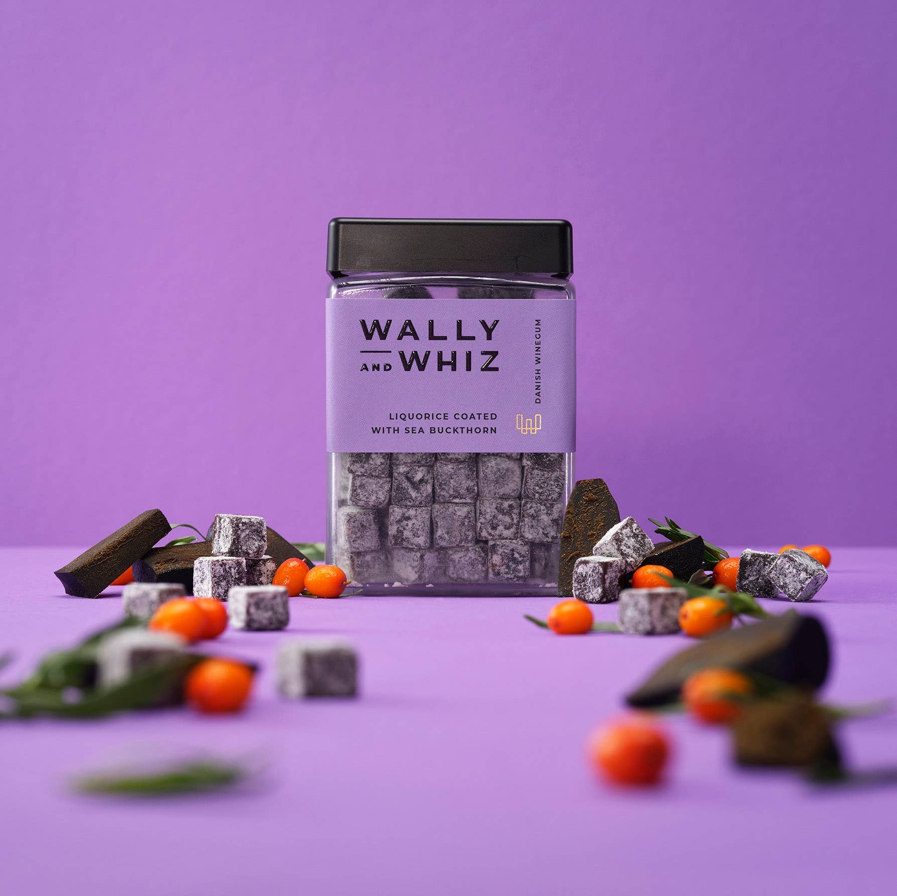 A jar of Wally & Whiz - Danish Winegum Candy Gummies sits on a purple surface, surrounded by licorice cubes, sea buckthorn berries, and green leaves against a vibrant purple background.