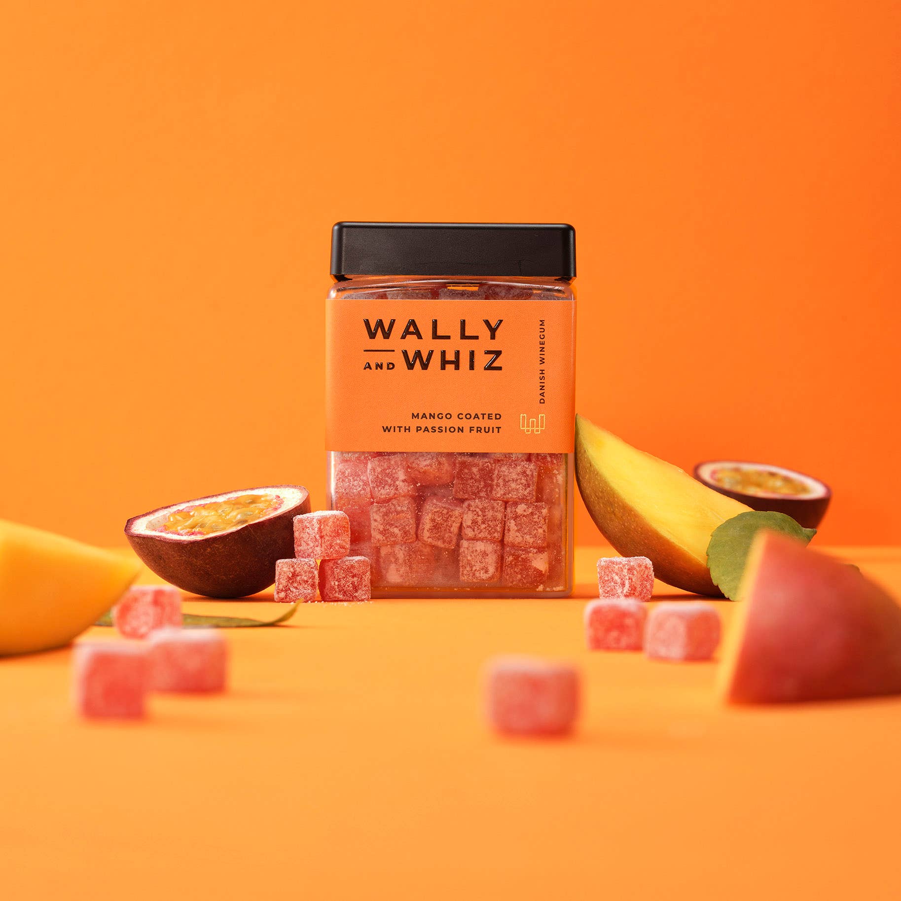 A jar of Wally & Whiz - Danish Winegum Candy Gummies in mango and passion fruit flavors is surrounded by fruit pieces and sugar-coated vegan gummies against an orange background.