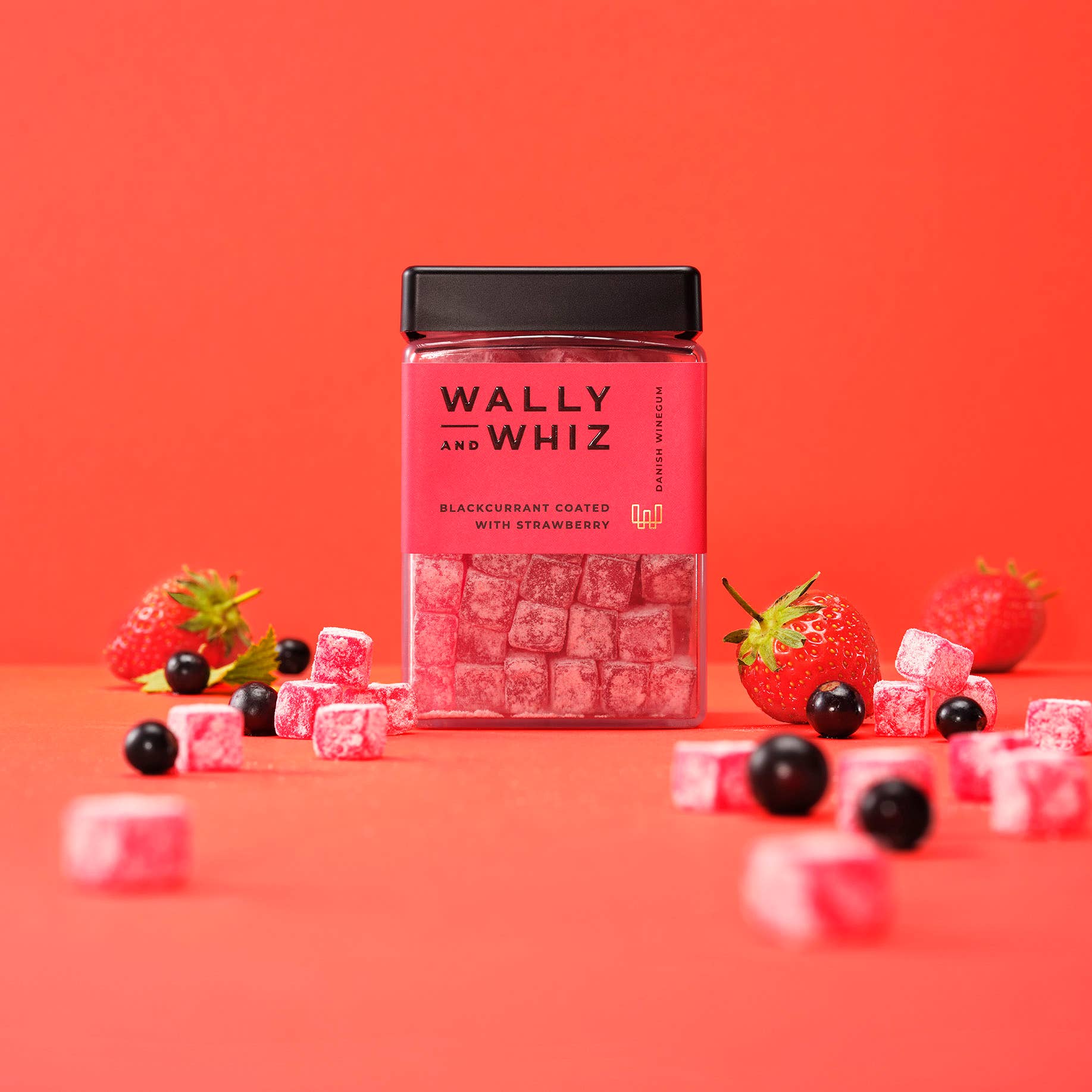 A jar of Wally & Whiz - Danish Winegum Candy Gummies—blackcurrant coated with strawberry—rests among scattered vegan gummies, strawberries, and blackcurrants on a red background.