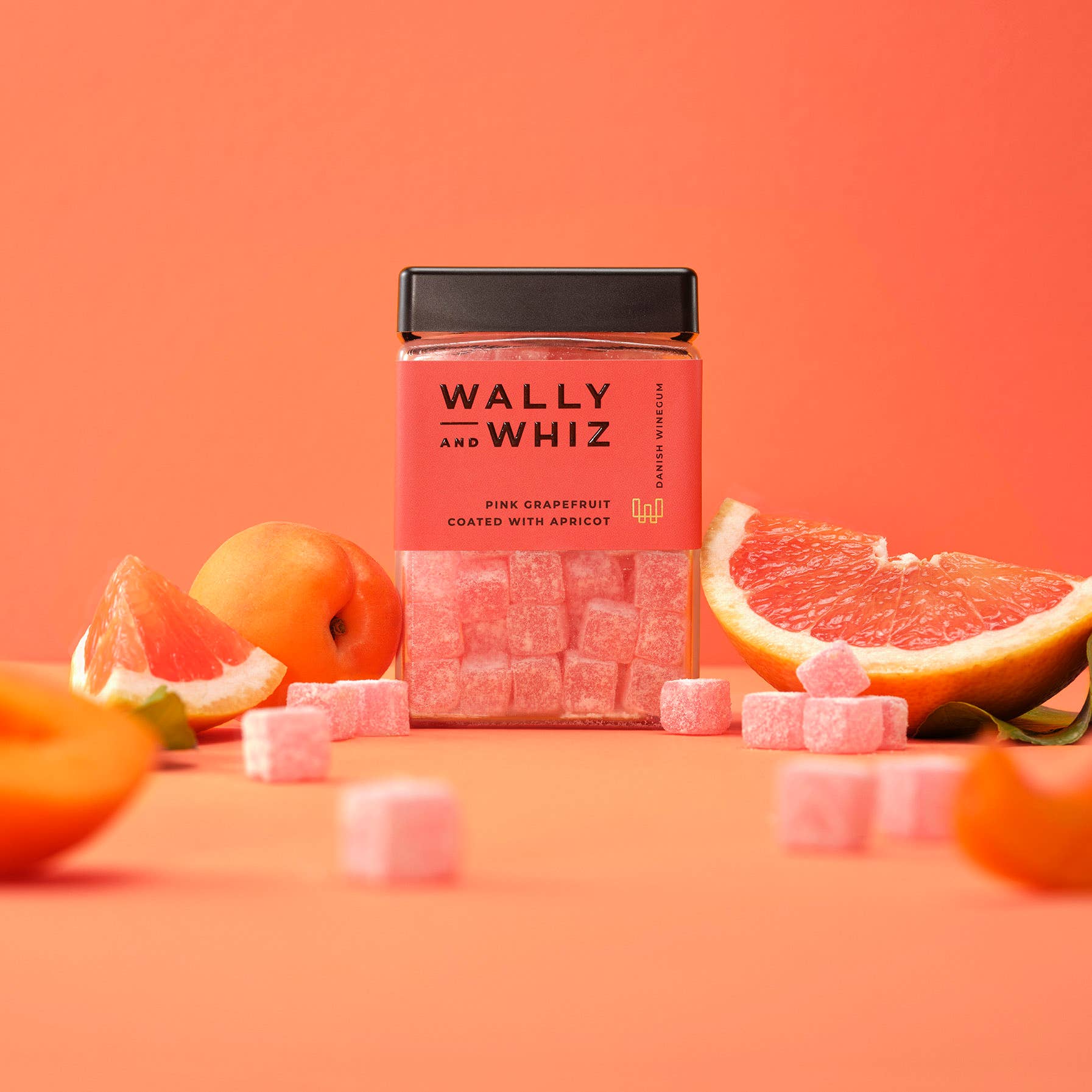 A jar of Wally & Whiz - Danish Winegum Candy Gummies in pink grapefruit and apricot flavor sits among fresh apricot halves and grapefruit slices on a peach-colored background.