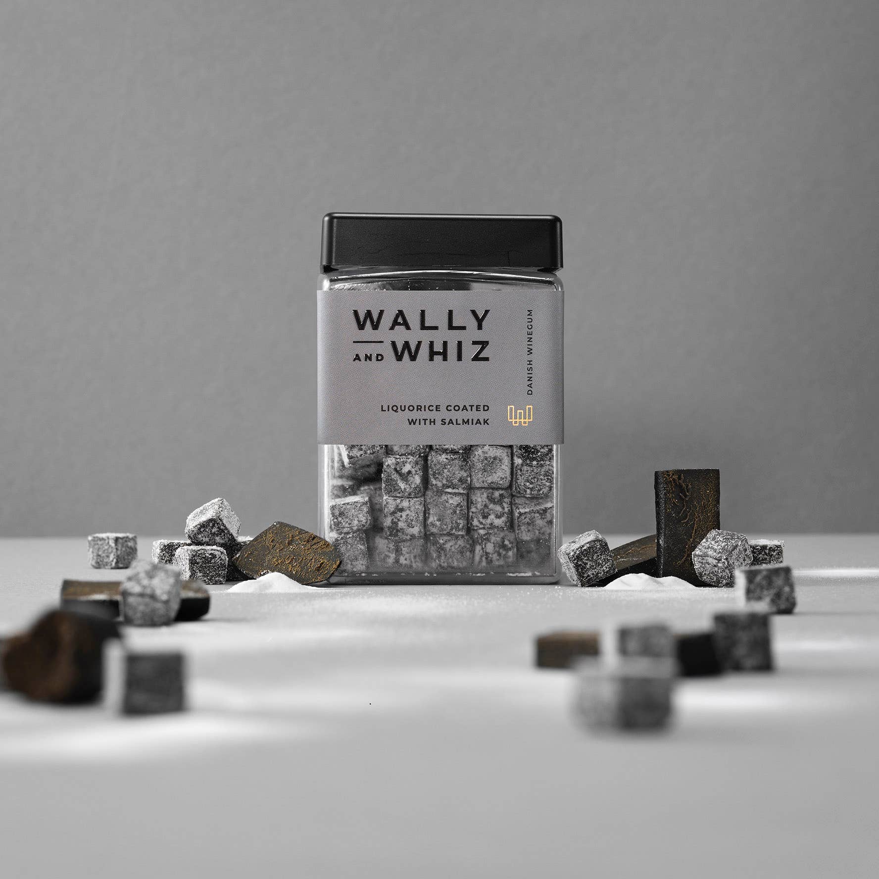 A clear jar labeled Wally & Whiz - Danish Winegum Candy Gummies holds liquorice-flavored cubes, with extra gummies scattered on a white surface against a grey background.