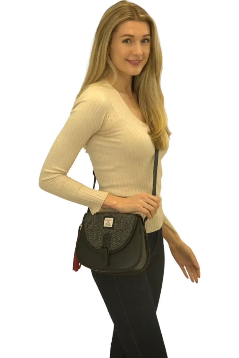 Woman with long blonde hair in a beige ribbed sweater and dark pants holds the Harris Tweed Saddle Bag in Grey, standing against a white background.