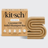 The KITSCH Coconut Oil Shampoo Bar for Dry Damaged Hair (3.2 oz) is uniquely designed in an abstract “S” shape and comes next to its tan packaging. Specially labeled as a moisturizing shampoo for kinky, curly, and coily hair, this solid bar highlights icons for bio-based ingredients, USA-made quality, cruelty-free standards, and sustainable sourcing. Infused with nourishing coconut oil, it helps enhance and care for your natural curl pattern.
