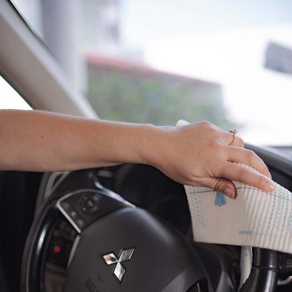 A person wipes a car steering wheel with the "Life is Better at the Lake" Swedish dish cloth, ensuring an eco-friendly clean.