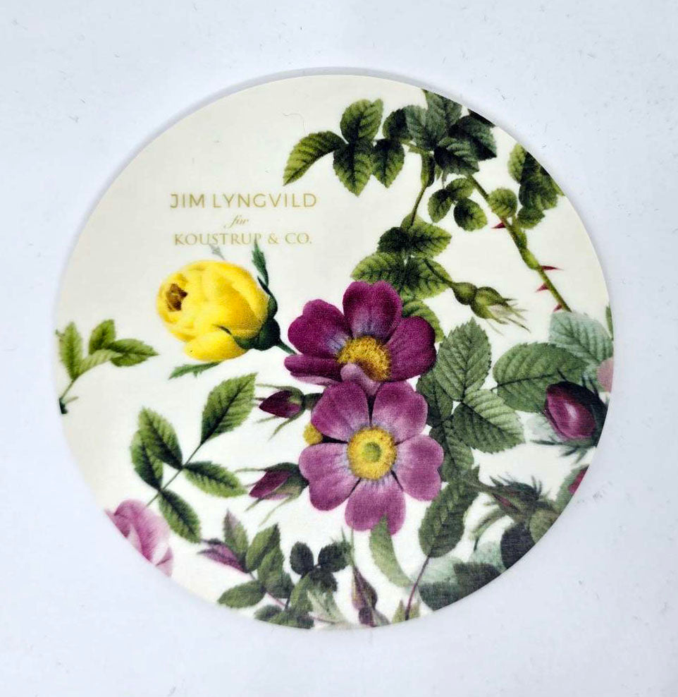 A round Nordic birch veneer coaster featuring an illustration of purple and yellow flowers, reminiscent of a rose flower garden, beautifully intertwined with green leaves. The text states "Danish designer Jim Lyngvild for Koustrup & Co.