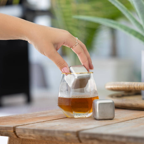 A hand places a Cubes: Stainless Steel Ice Cube (1.57" square) into a glass of amber liquid, while another reusable cube from the set of 2 rests in its plastic case on the wooden table nearby.