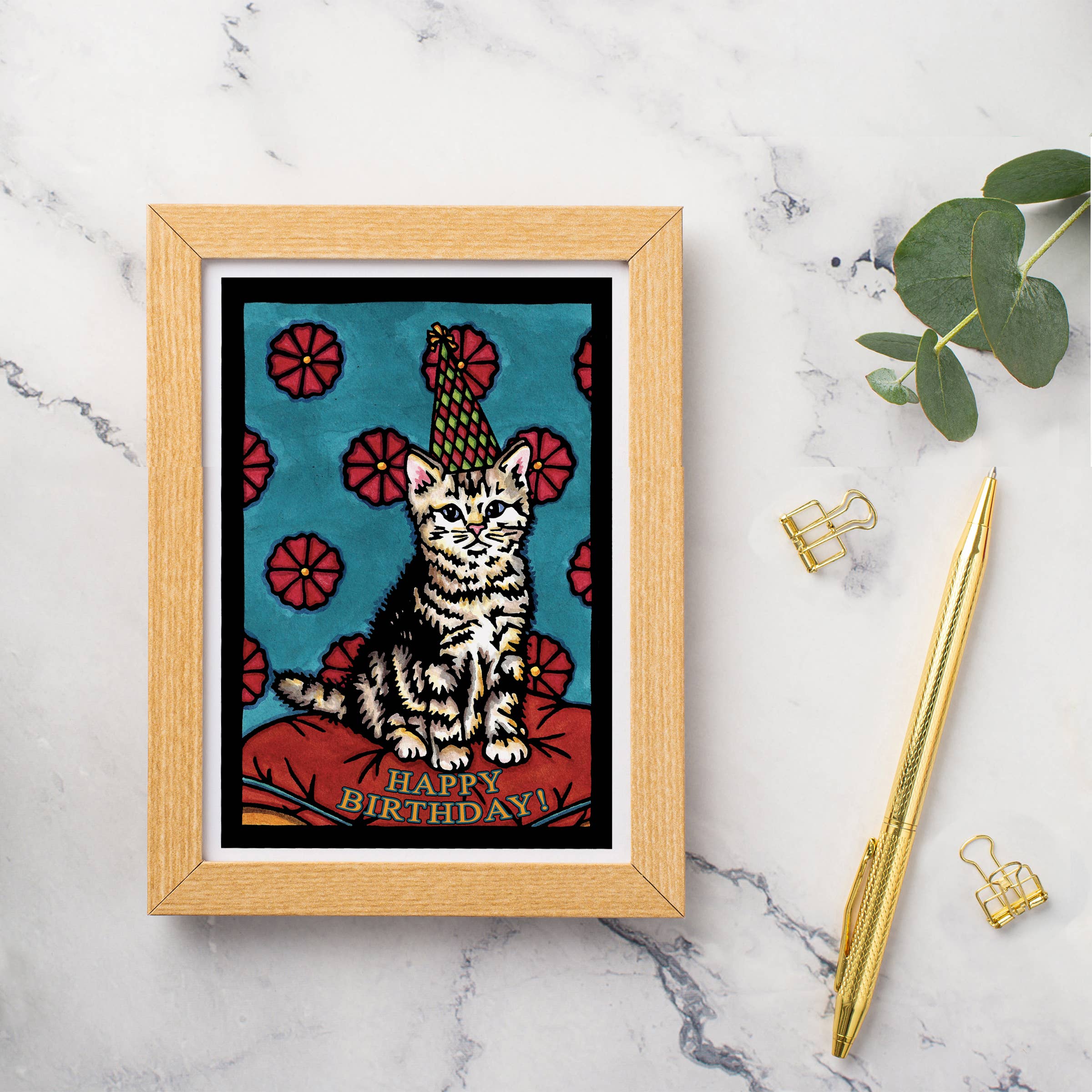 The "Birthday Kitten" greeting card by Sarah Angst, featuring a cat adorned with a party hat and the text "Happy Birthday!" captures the whimsical charm typical of her art. It is elegantly displayed on a marble surface surrounded by a gold pen, paper clips, and green leaves.