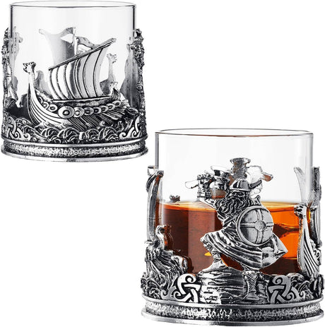 The Viking & Mythological Whiskey Drinking Glass (12 OZ) features two clear glasses with ornate silver bases, showcasing ship and warrior motifs; one glass is filled with amber old fashioned liquor.