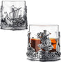 The Viking & Mythological Whiskey Drinking Glass (12 OZ) features two clear glasses with ornate silver bases, showcasing ship and warrior motifs; one glass is filled with amber old fashioned liquor.
