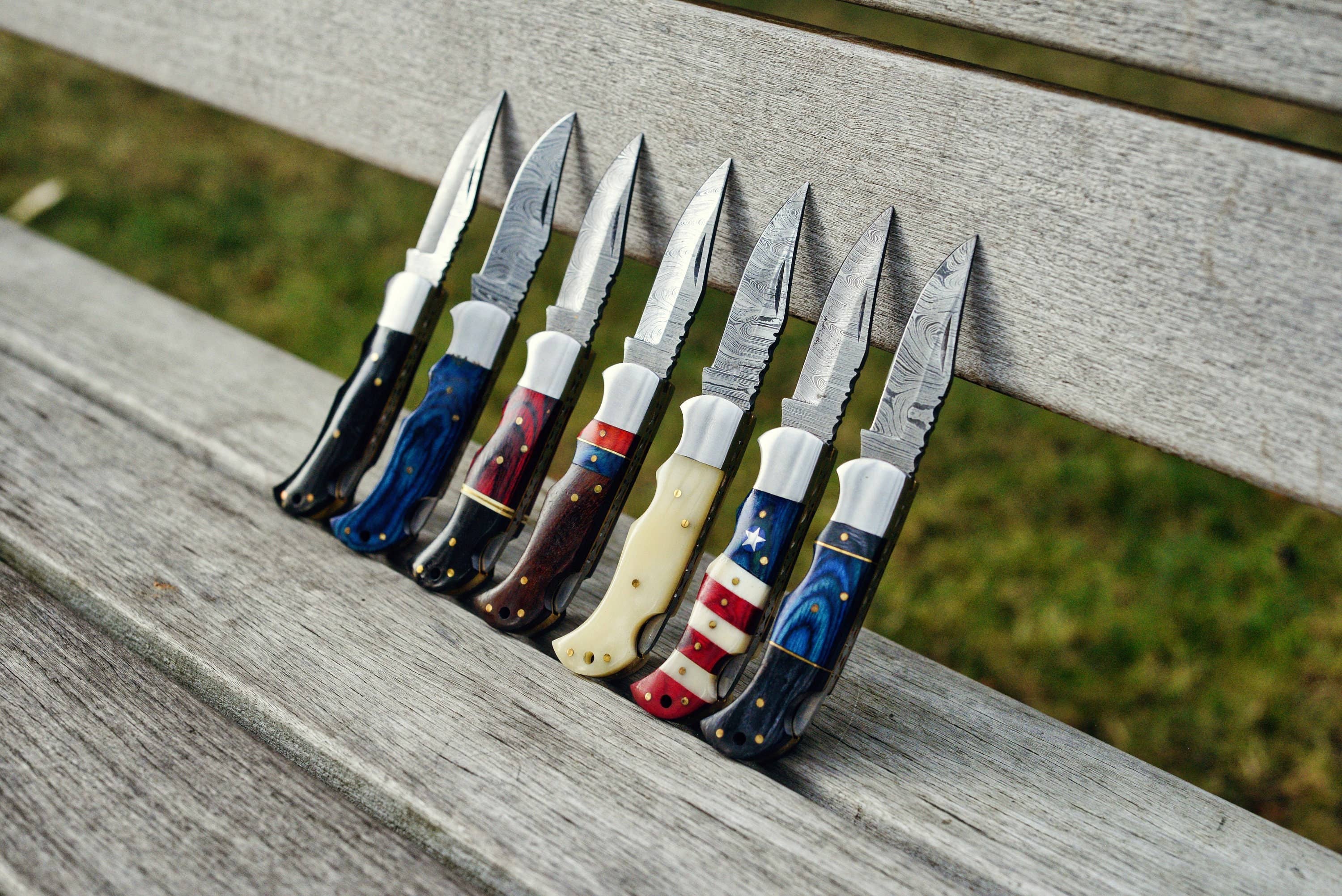 Seven Blue Damascus folding knives with decorative blue wood handles are lined up on a wooden bench.