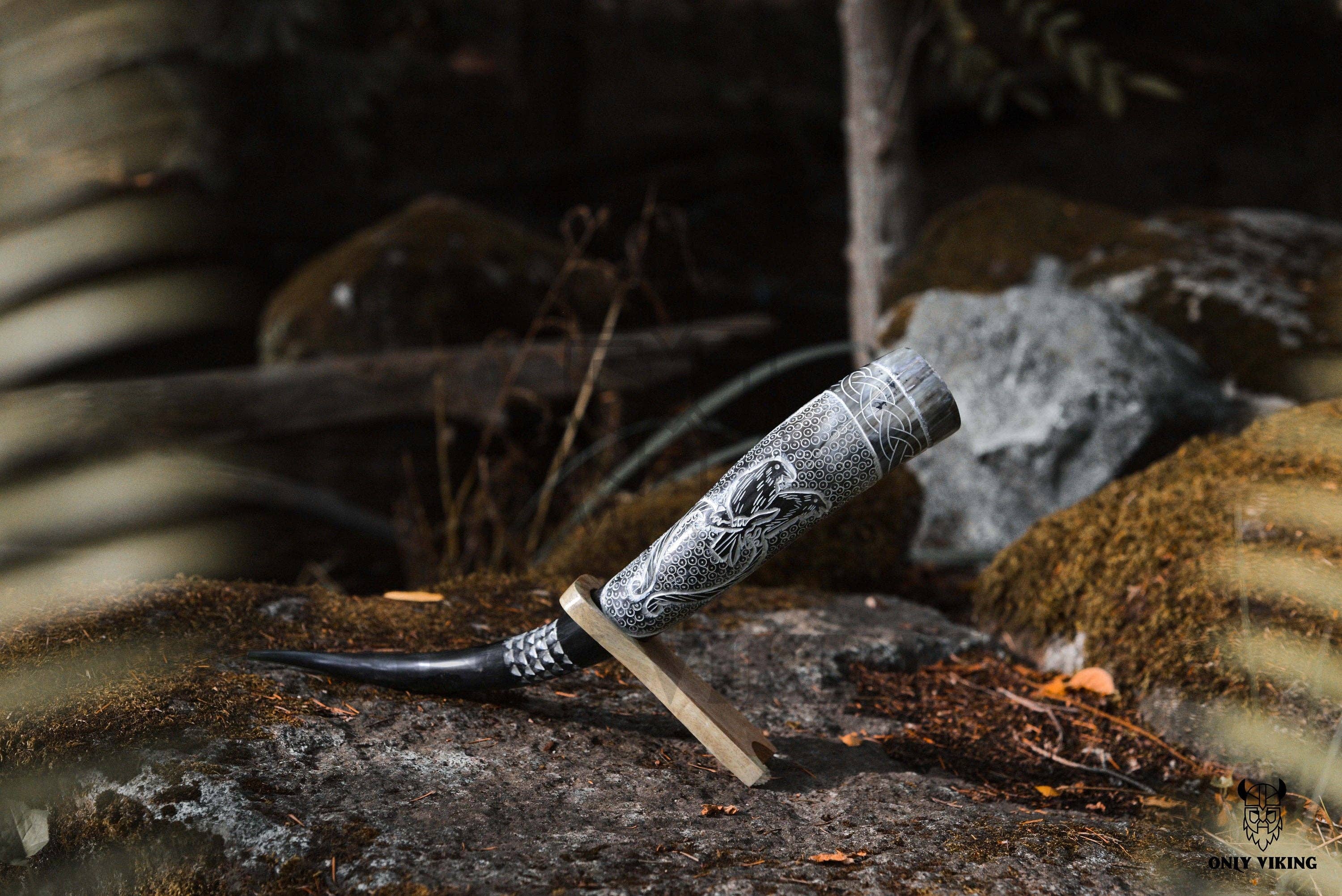 The Ravens Viking Ship Handcrafted Drinking Horn (14-18) with metal stand is displayed outdoors among rocks and moss.