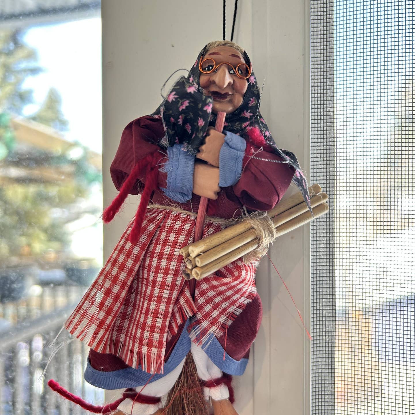 The Decor: Kitchen Witch Doll Opalina Red 8.3" Tall features a headscarf, glasses, plaid apron, red dress, and bundle of sticks—perfect as a traditional good luck charm to hang in your home.
