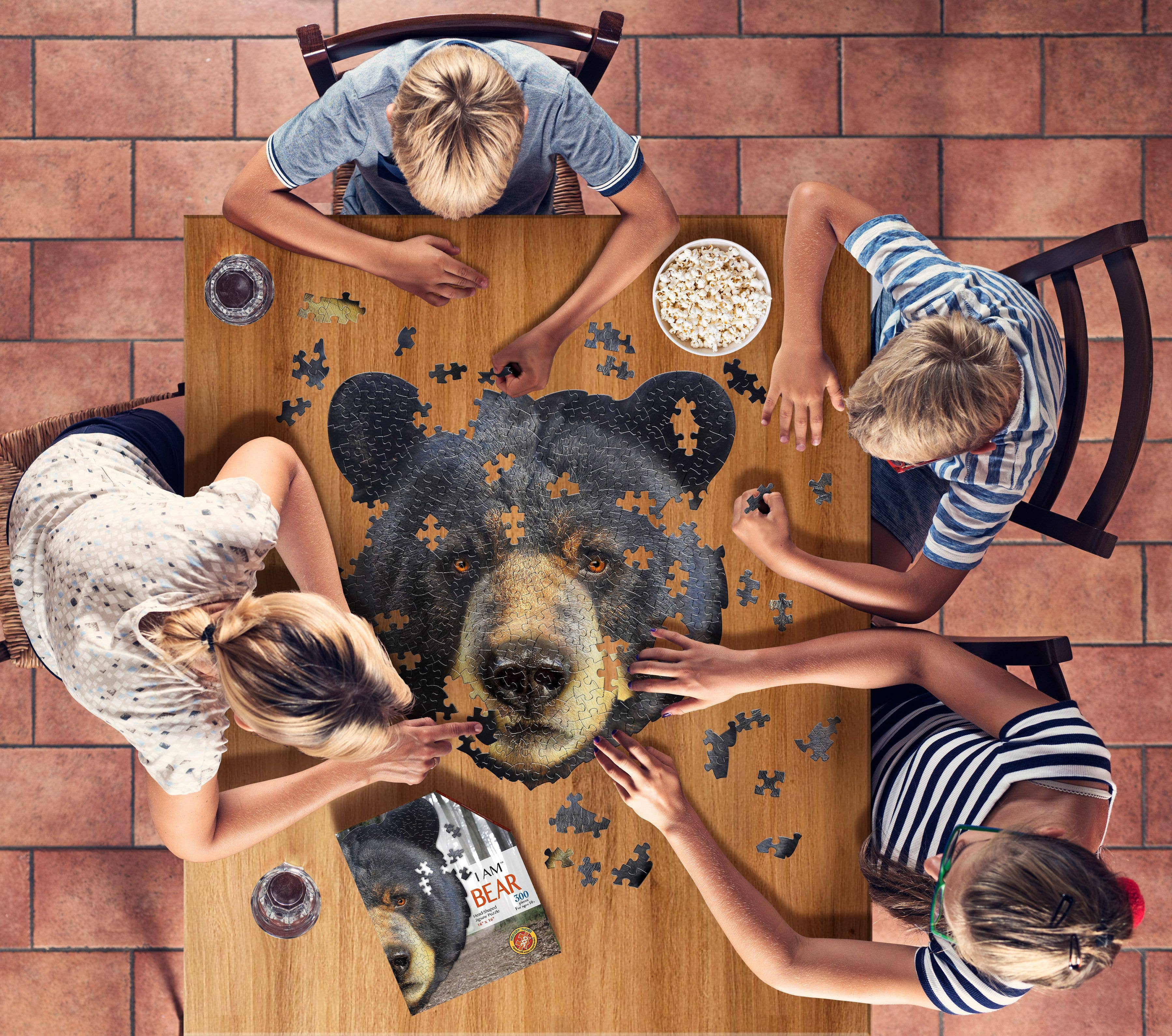 Top view of four people working on the I Am Bear-shaped jigsaw puzzle (300 pieces) on a wooden table, with a bowl of popcorn and the puzzle box nearby.