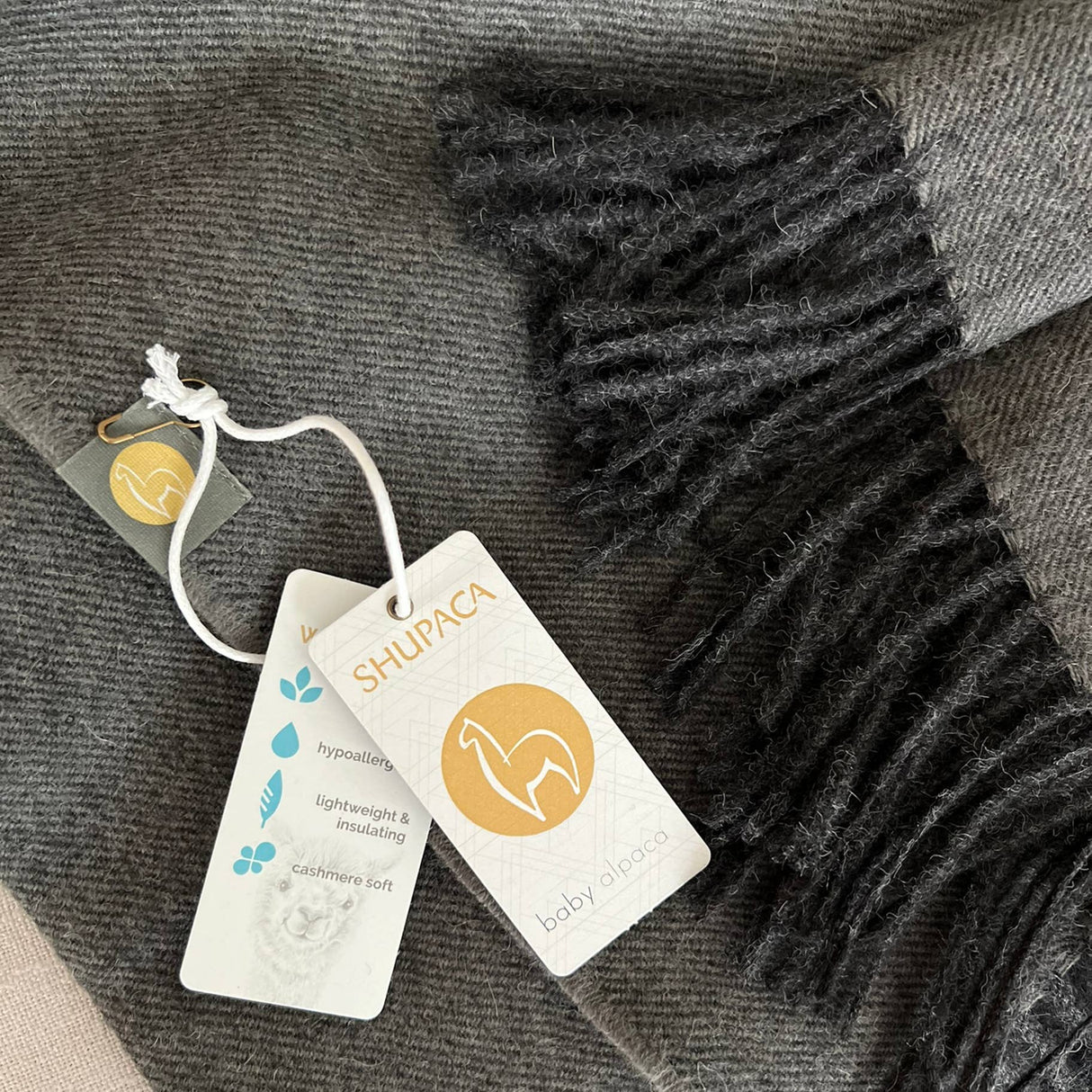 A unisex 100% Baby Alpaca Scarf in Carbon (charcoal) is shown with fringed ends and attached tags labeled SHUPACA, baby alpaca, and showing product features and logo.