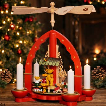 A handcrafted wooden Christmas pyramid made in Germany features spinning blades on top and festive figurines—including a bear and other animals—set amidst Christmas lights and pinecones, and is illuminated by Marketplace24 White Christmas Pyramid Candles (Pyramiden-Kerzen), available in a box of 50 candles (14 x 70mm).
