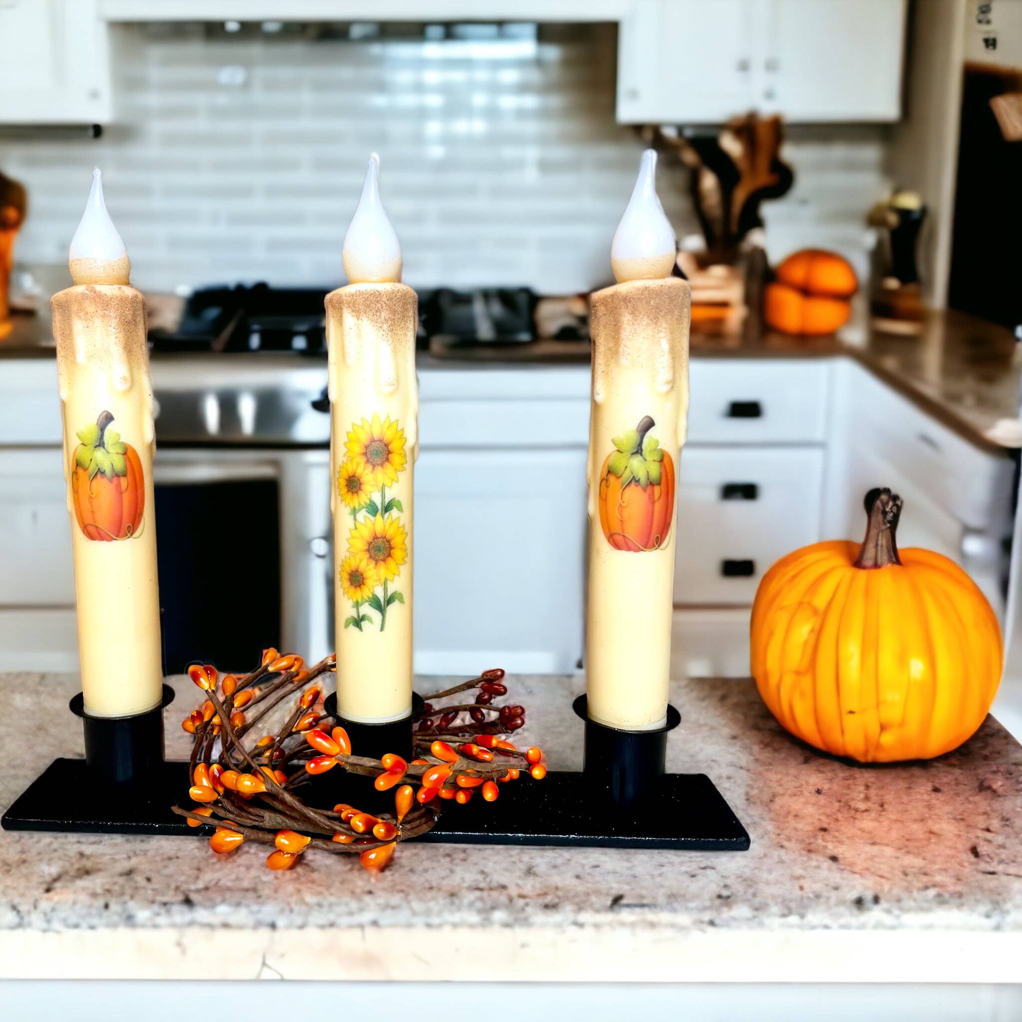 A cozy centerpiece in the kitchen features three decorative candles adorned with pumpkin and sunflower motifs, nestled in the Triple Wrought Iron Taper Holder on a counter, accompanied by a small pumpkin and an orange berry garland.