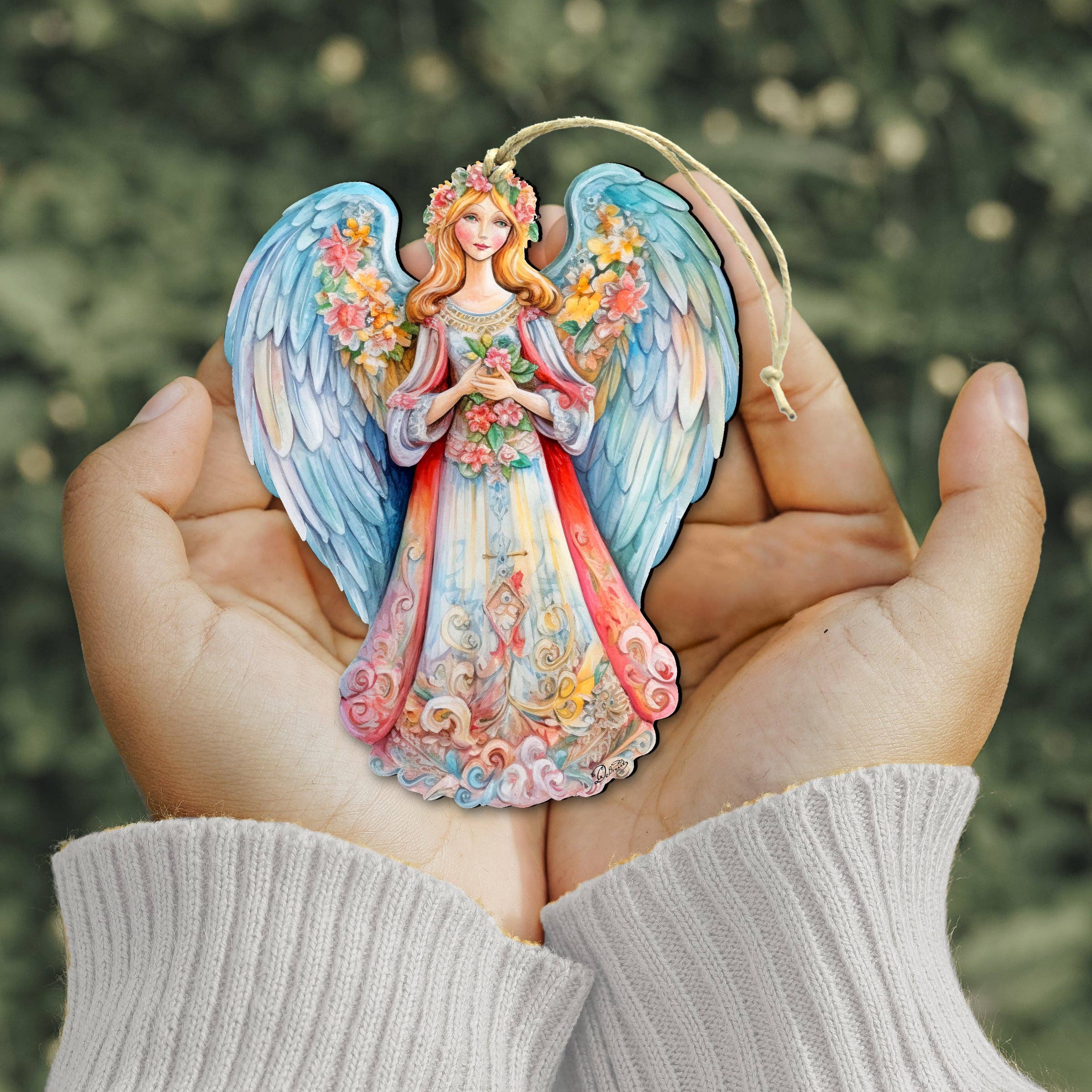 In softly cupped hands, a beautifully crafted keepsake is held—a vibrant angel ornament from the Debrekht Nativity Holiday collection, featuring floral details and striking blue wings—set against a blurred green backdrop.