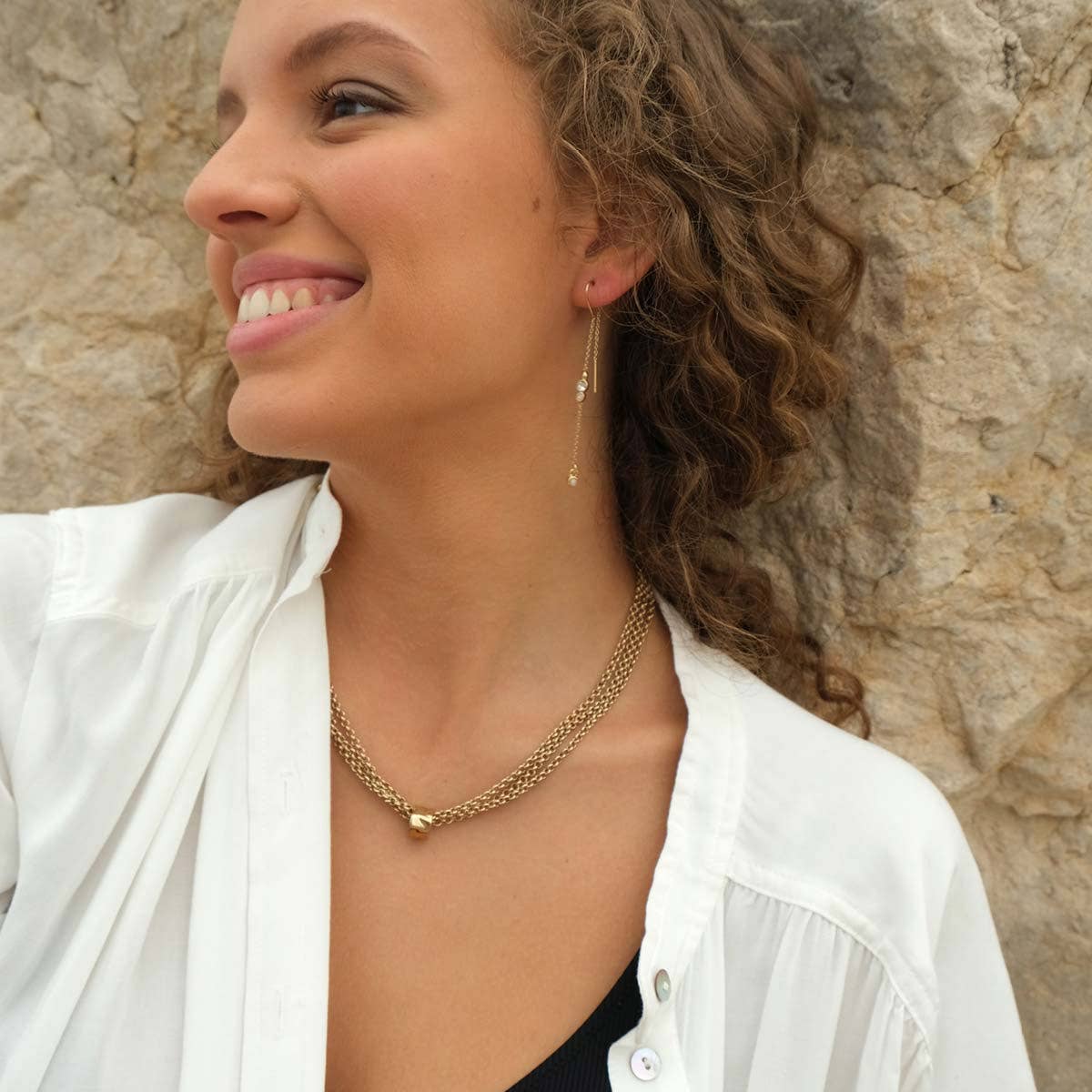 Woman with curly hair wearing a white shirt, layered gold necklace, and A&C Oslo Long Crystal Chain Gold Earrings, smiling and looking to the side against a rocky background.