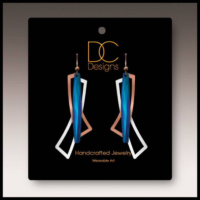 A pair of modern blue, silver, and copper geometric open wings earrings (1.75"), handmade in stainless steel, displayed on a black card labeled “DC Designs Handcrafted Jewelry Wearable Art.”.