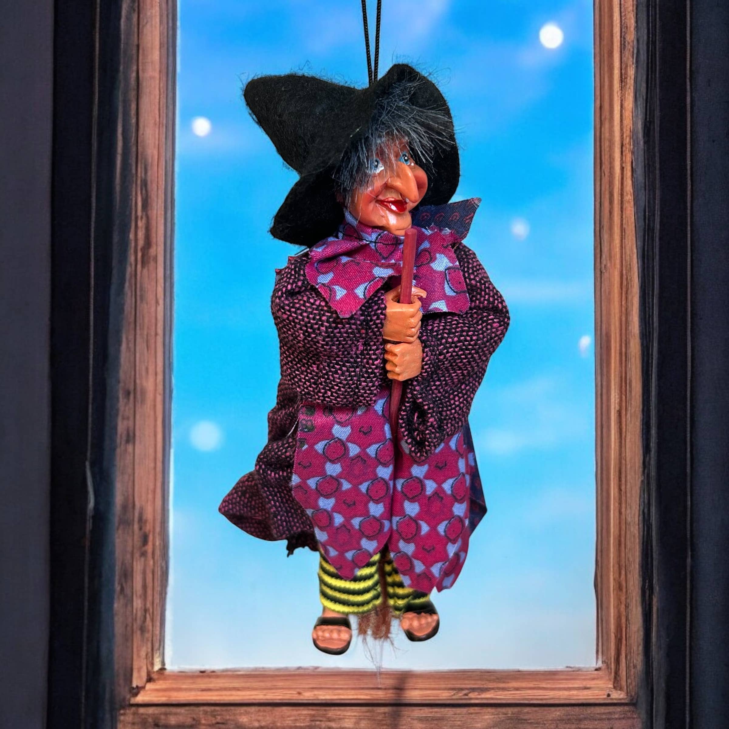 The Johanna Purple Kitchen Witch Doll Figurine, 9" tall, wears a black hat and purple dress, hanging by a string in front of a window against a blue sky—a whimsical good luck charm for any home.