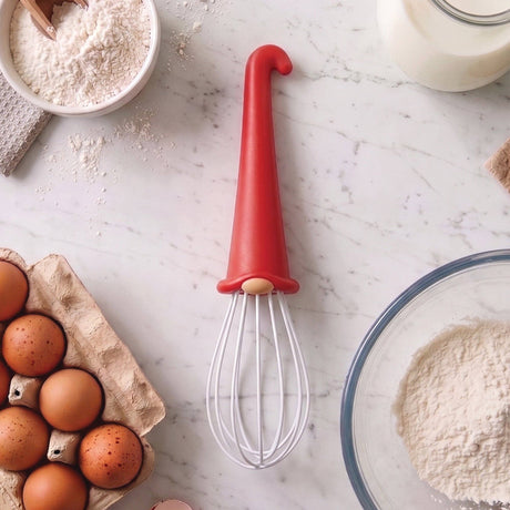A playful OTOTO Whisk: Gnome Wire Whisk, featuring a vibrant red handle shaped like a gnome's hat, rests on a marble countertop amidst eggs, flour, and a glass of milk—setting the scene for baking fun. This charming kitchen tool brings joy and whimsy to any kitchen and makes an adorable gift for any home baker. When batter needs blending & cream needs a whip, he spins through the bowl with a hop & a skip!.