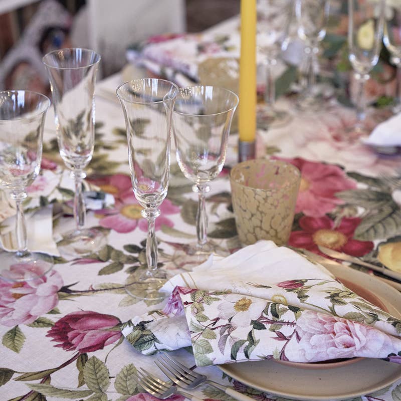 A table set with a floral tablecloth, napkins, wine glasses, a yellow candle, beige plate, and the Rose Flower Garden Round Serving Tray 14.9 by Koustrupco of Denmark brings Nordic charm to your dining experience.