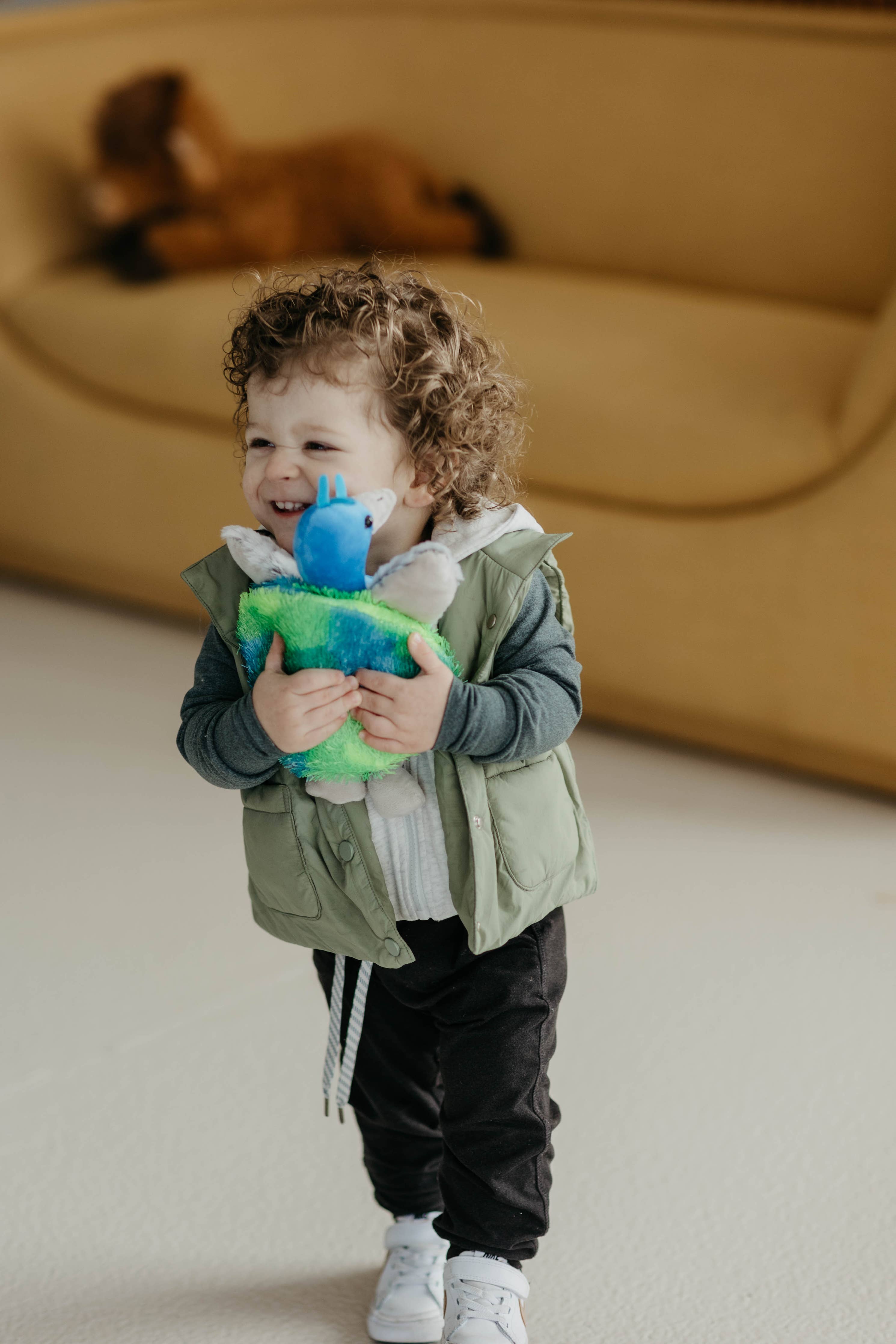 A young child with curly hair smiles while hugging the Wild Onez Plush Peacock Stuffed Animal, made from recycled water bottle fiber, in a room with a tan sofa in the background.