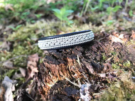The Lapland Sami Bracelet #2—crafted from reindeer hyde with pewter and silver bands in black—rests on a mossy tree stump in a wooded outdoor setting.