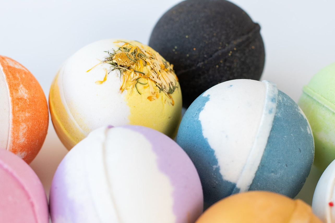 A close-up of several colorful handmade bath bombs, including the Bath Bomb: Sandalwood & Vanilla variety in shades like white, yellow, blue, purple, black, green, pink, and orange—each with unique textures and nourishing ingredients.