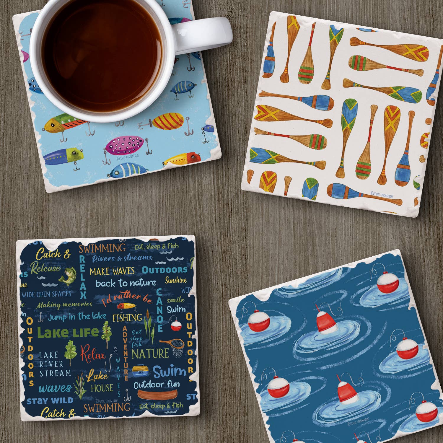 Resting on a wooden surface are four vibrant coasters with a tumbled tile edge, showcasing designs of fish, paddles, fishing bobbers, and words inspired by the outdoors. Each Lake Living Coaster #3 is crafted from absorbent stone and includes a cork backing for protection. A cup of coffee is placed on the coaster adorned with the fish design.