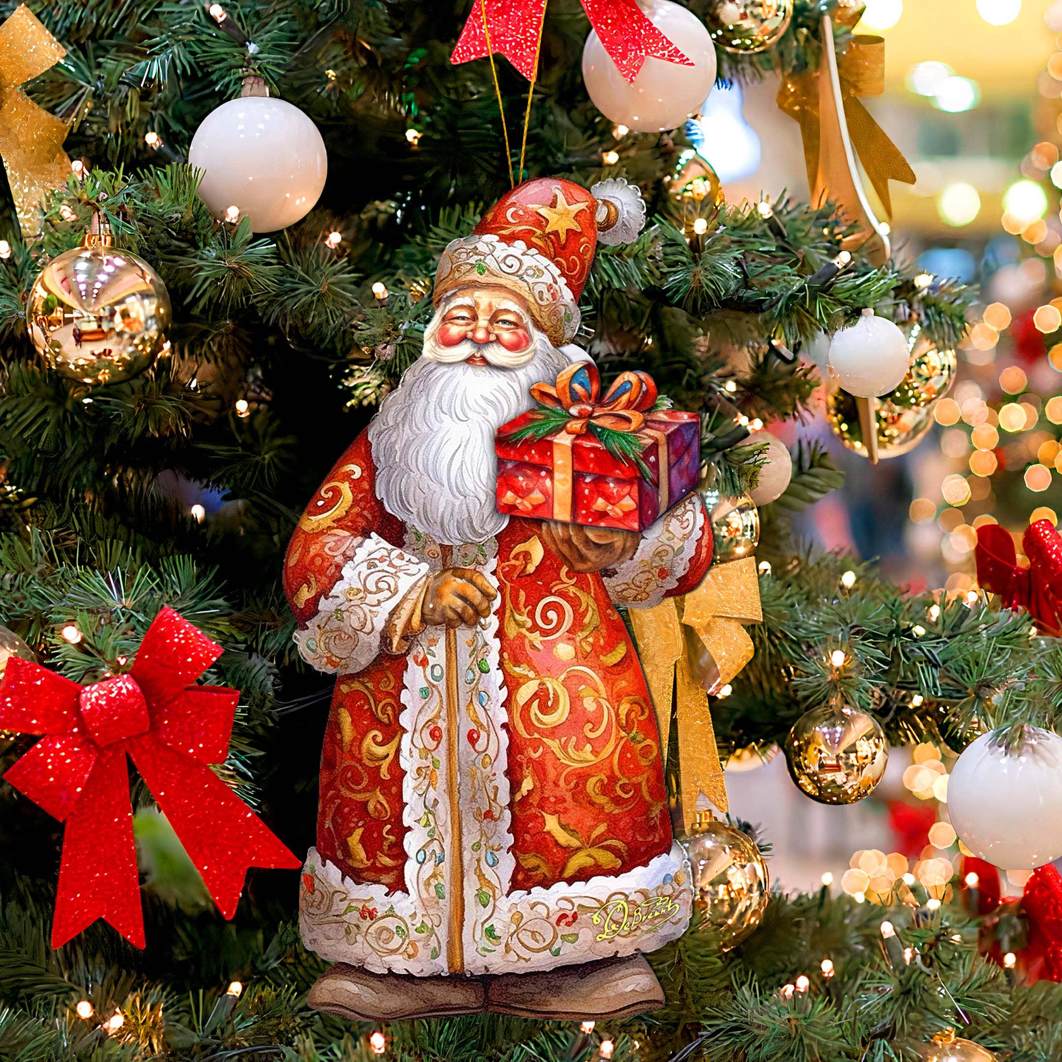 A handcrafted "Ornament: St. Nick's Gift Wood Ornaments GDebrekht Christmas Santa" hangs gracefully on a decorated Christmas tree adorned with red bows and various ornaments, adding to the festive holiday décor with its depiction of Santa Claus holding a gift and dressed in red and gold.