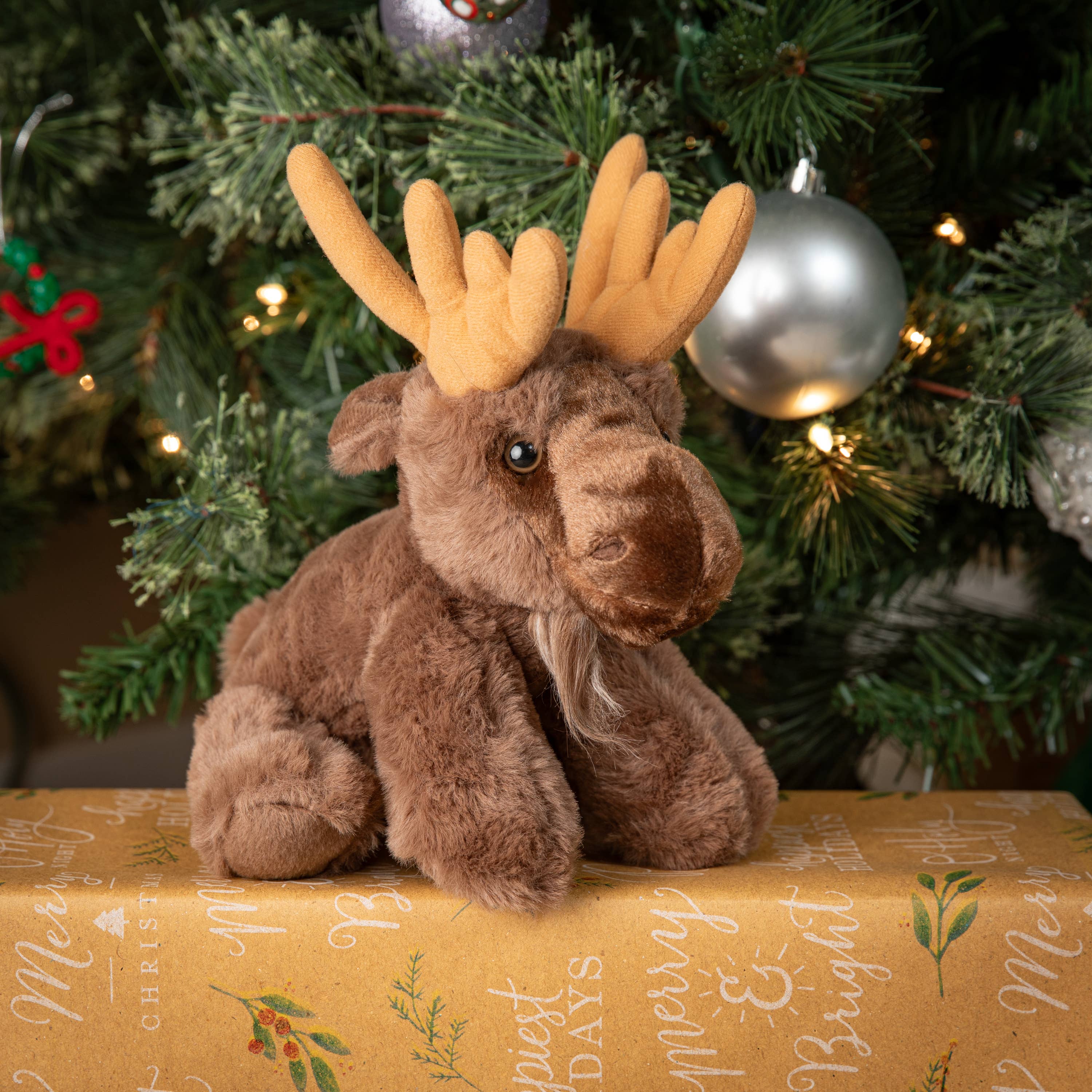 A Plush: Moose 9 Tall Stuffed Animal Wild Onez sits atop a wrapped gift box in front of a Christmas tree adorned with ornaments and lights.