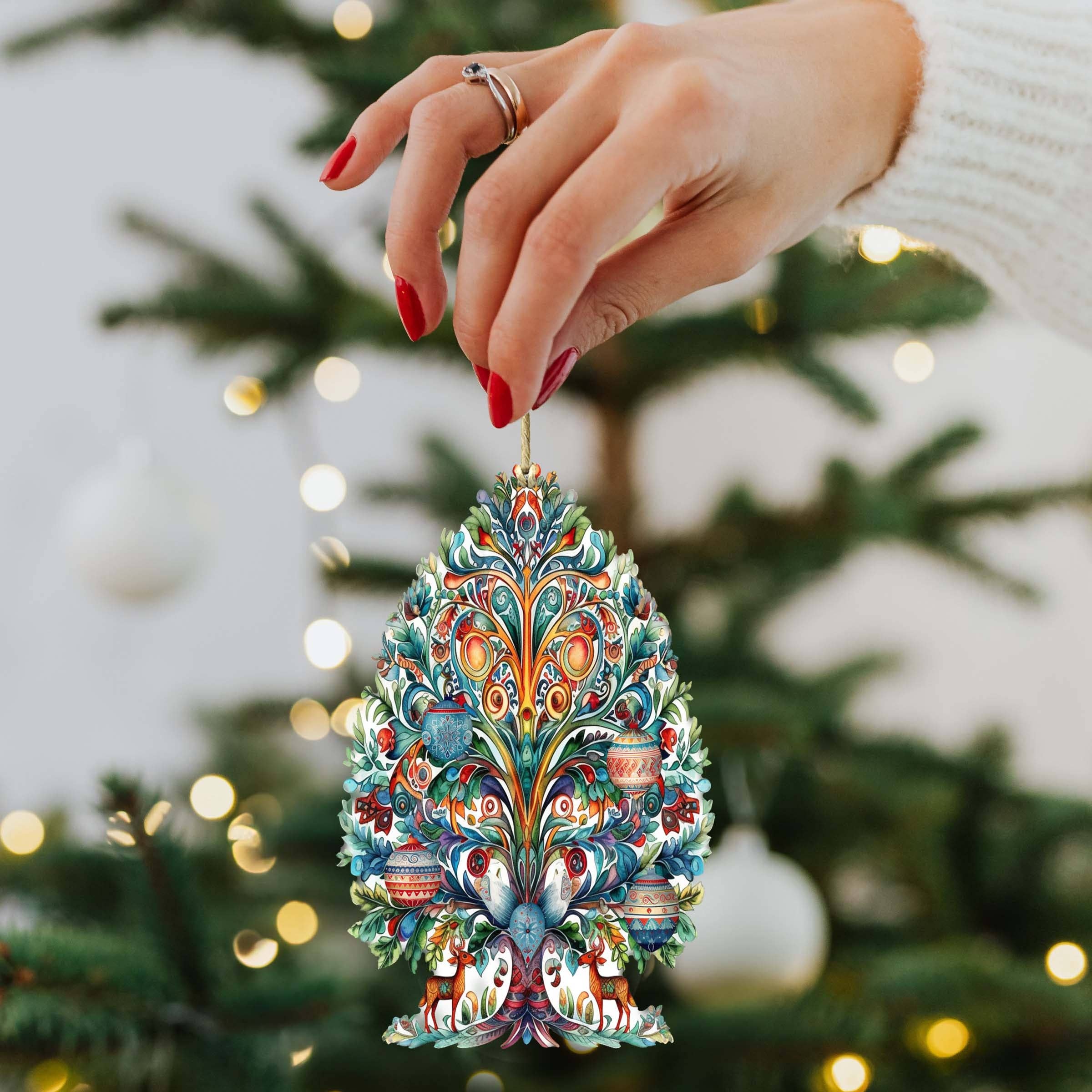 A hand with red nails holds a vibrant Nordic Christmas Tree wooden ornament, beautifully handcrafted, in front of a lit Christmas tree, enhancing the charm of the festive décor.