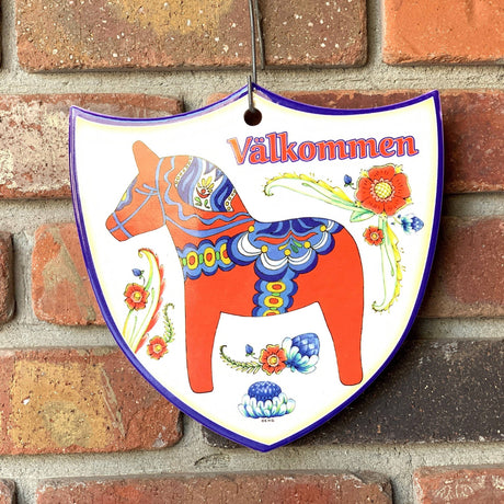 The Tile: Red Dala Horse Valkommen Ceramic Decoration Shield features a painted red Dala horse, colorful flowers, and Välkommen—a charming Swedish welcome sign perfect for home décor.