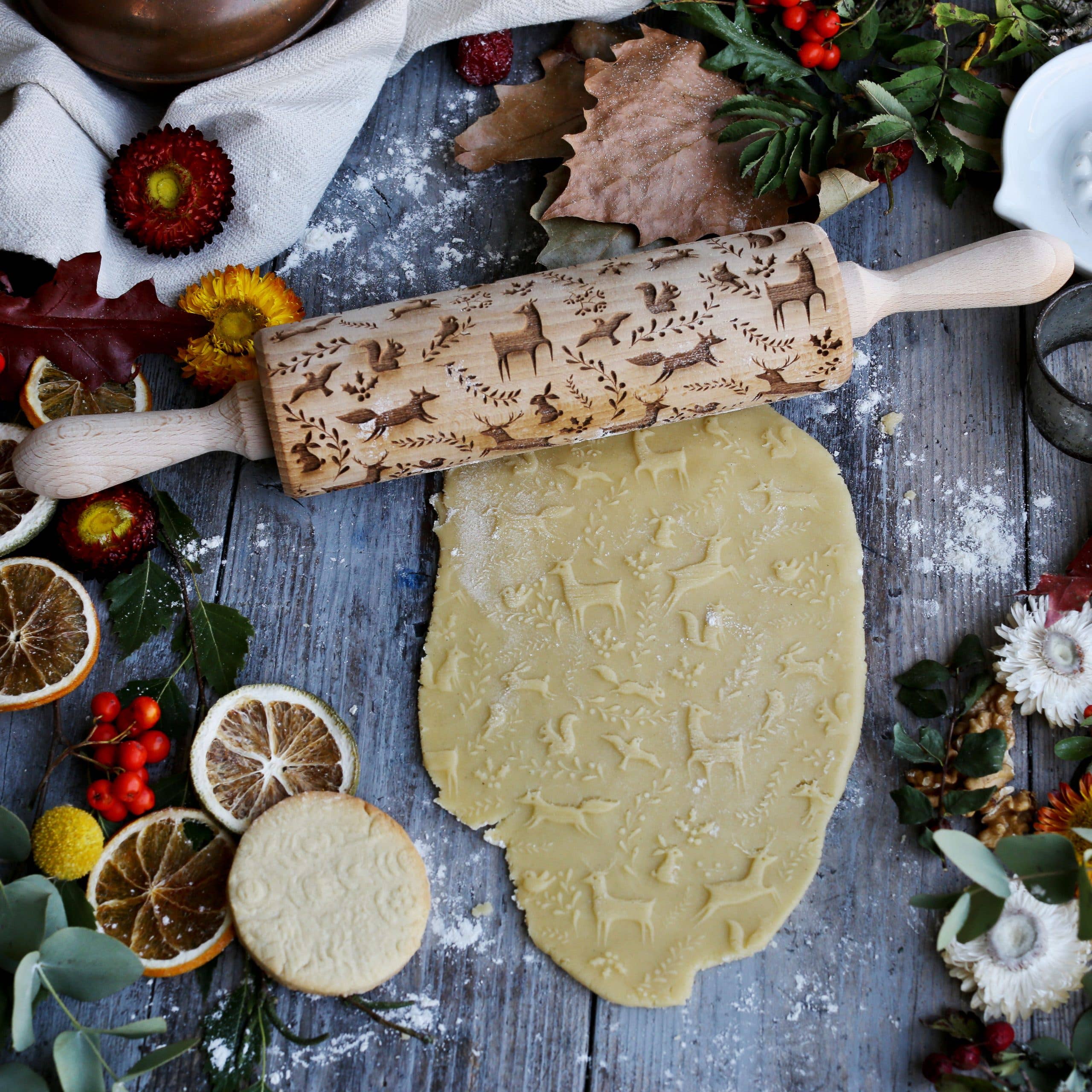 The Rolling Pin: Winter Woodland Animals Embossing Rolling Pin rolls out dough on a rustic table, surrounded by dried citrus slices, foliage, and flowers. This handmade item adds charm with its decorative engravings featuring winter woodland animals for a touch of nature.