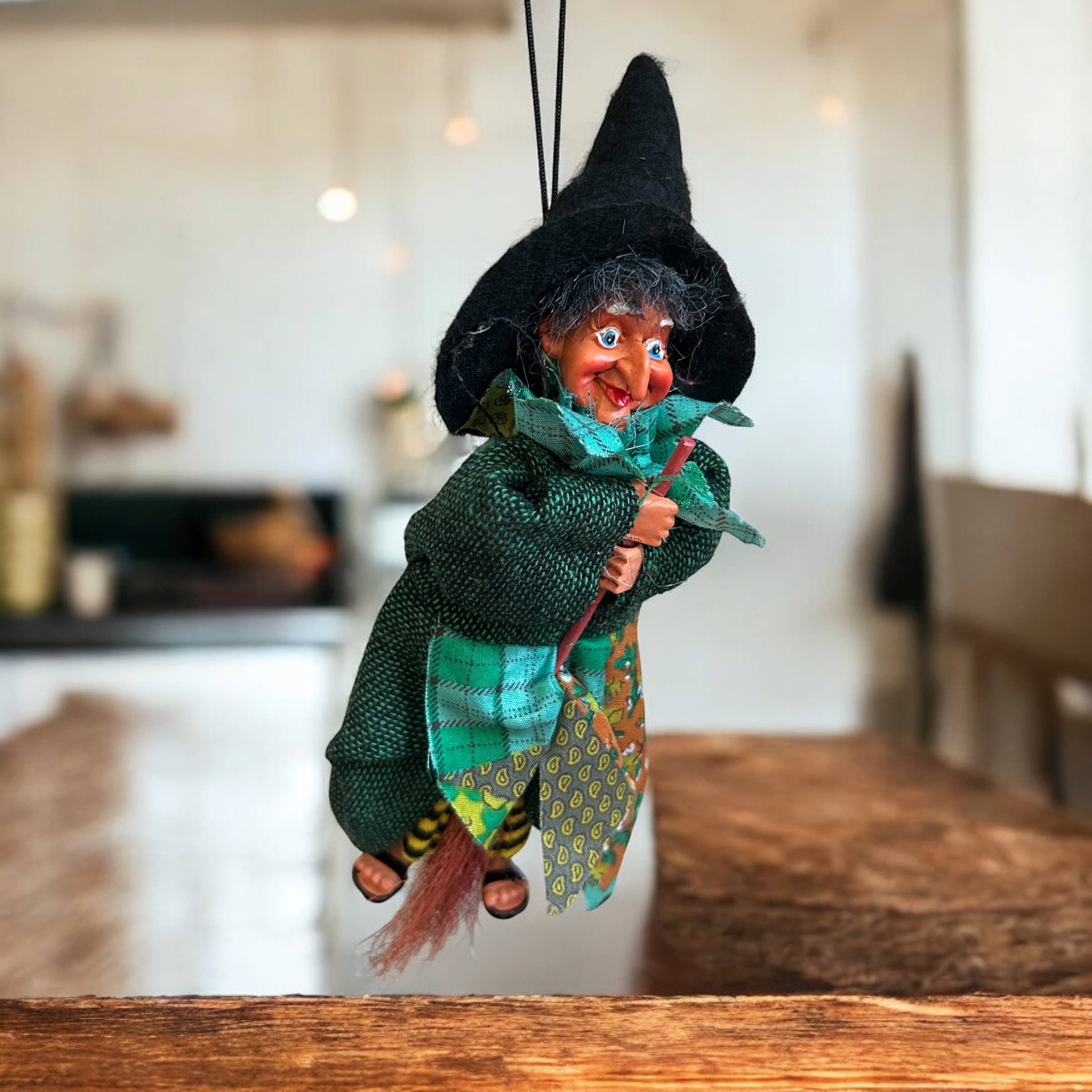 The Kitchen Witch: Johanna Green Kitchen Witch Doll, handmade in Germany, features a black hat, green clothes, and a broomstick, and hangs indoors by a string against a blurred background.