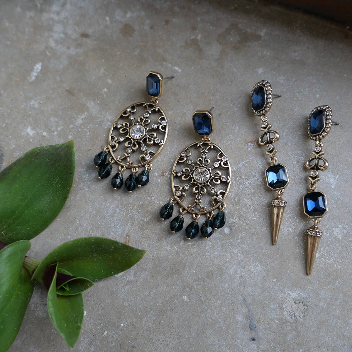 Two pairs of A&C Oslo Antoinette Midnight Blue Statement Earrings with ornate gold plating and blue gemstones are displayed on a stone surface beside a green plant.