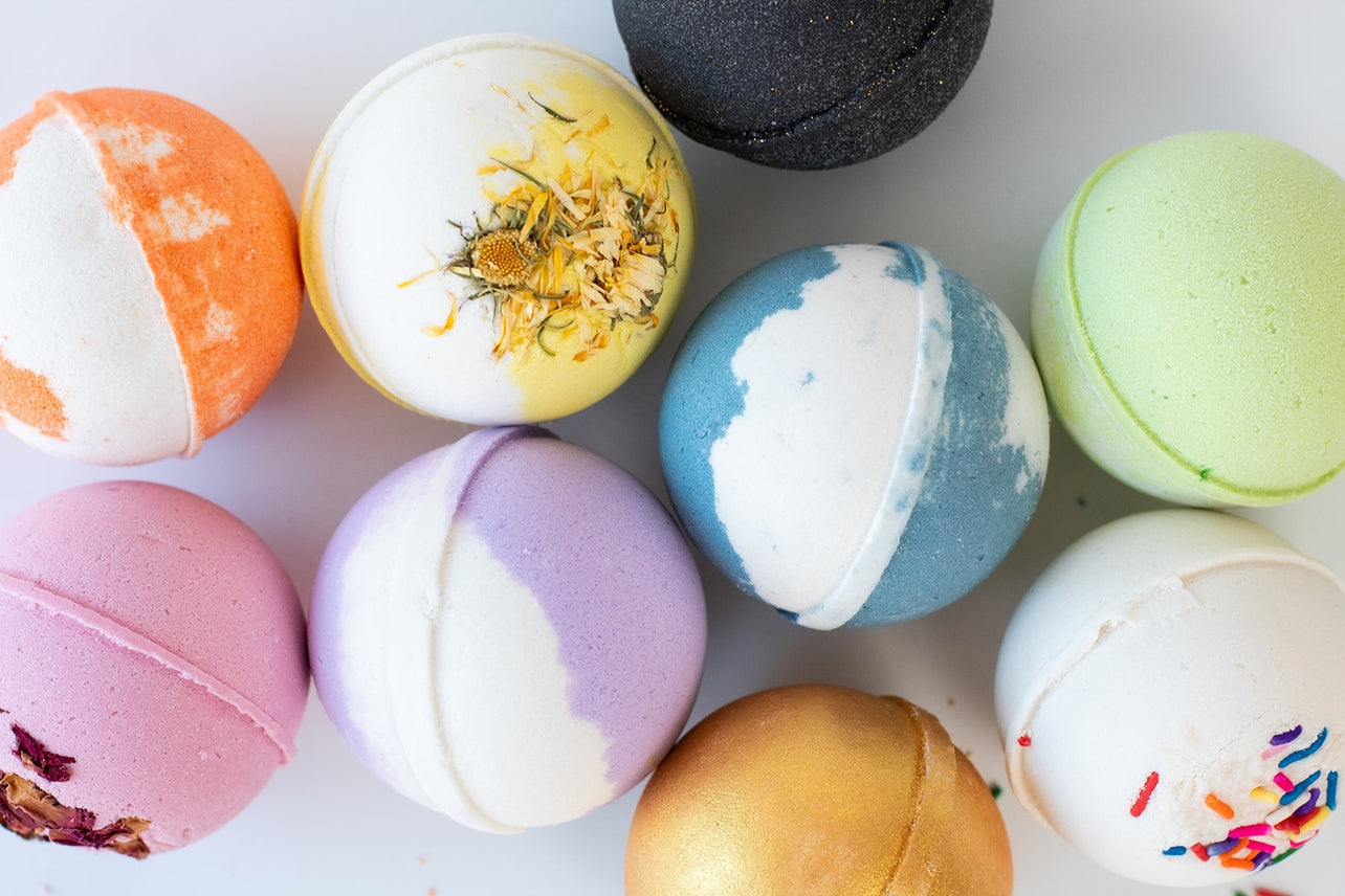 A variety of colorful handmade bath bombs, including the Sandalwood & Vanilla Bath Bomb with nourishing ingredients and decorative toppings, are arranged on a white surface.