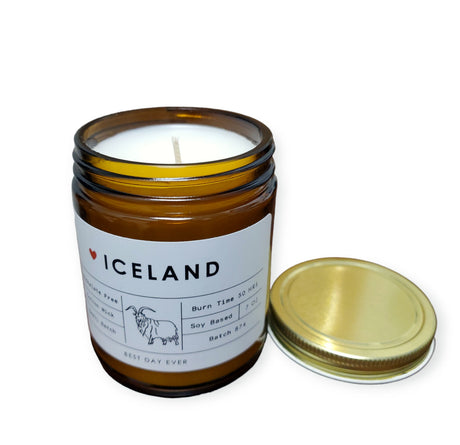 A brown glass Candle: Iceland Volcano Dupe Scent with a white label and gold metal lid off to the side, made from a soy coconut wax blend.