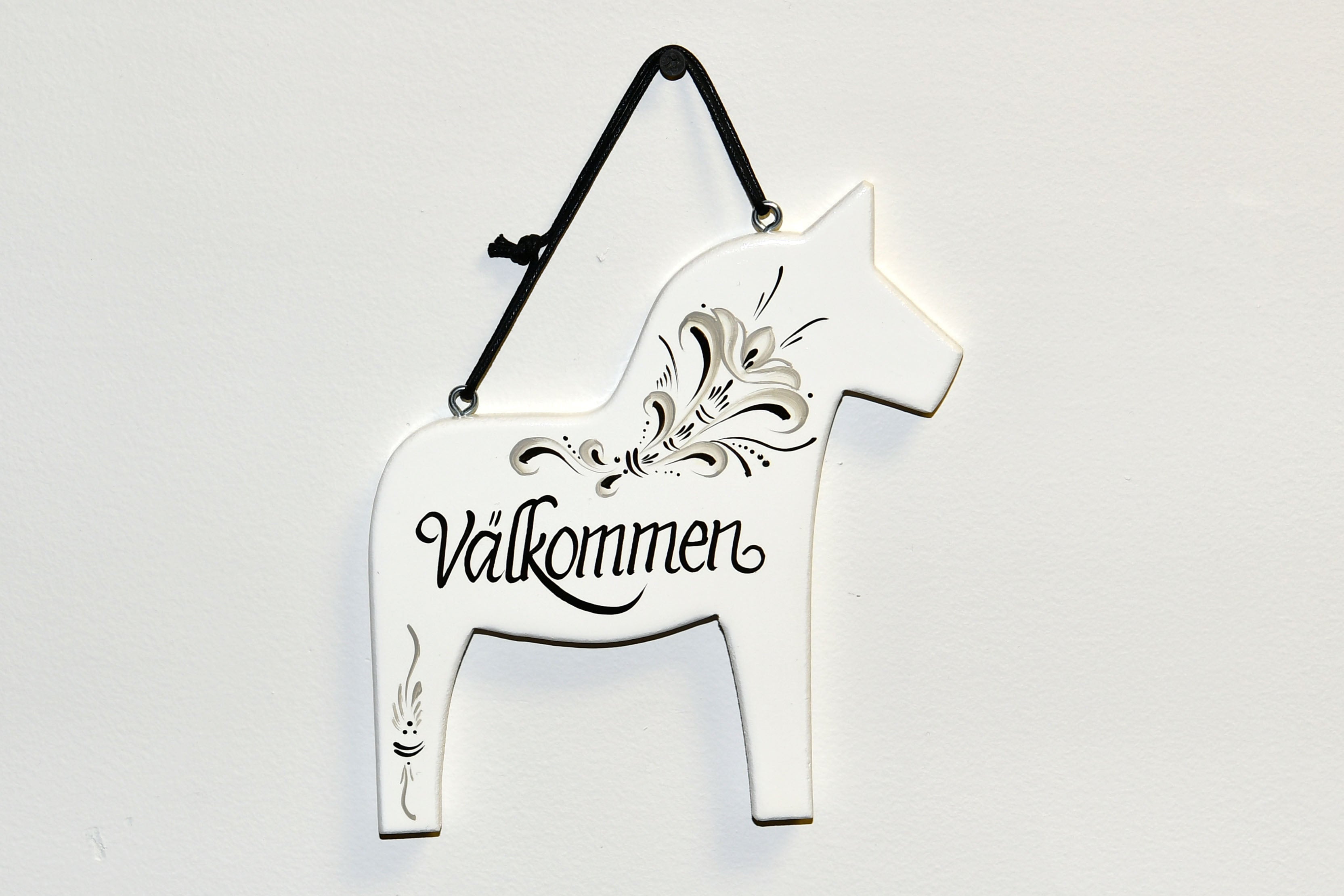 The White Swedish Dala Horse Välkommen Wall Decor features intricate black floral designs and the word Välkommen in cursive. It hangs elegantly from a black ribbon, bringing Scandinavian warmth to any space.