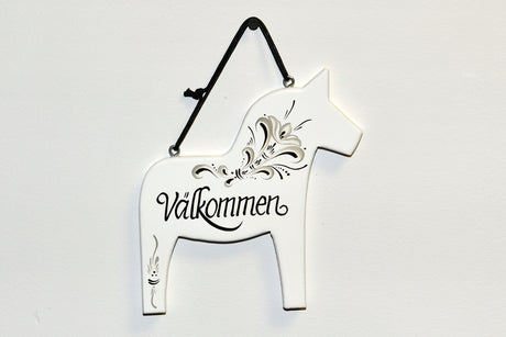 The White Swedish Dala Horse Välkommen Wall Decor features intricate black floral designs and the word Välkommen in cursive. It hangs elegantly from a black ribbon, bringing Scandinavian warmth to any space.