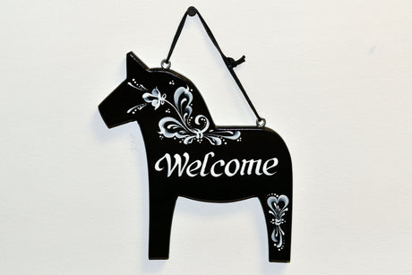 A black Swedish pine Dala horse-shaped Welcome wall decor features hand-painted white floral designs and hangs by a black cord against a white background.