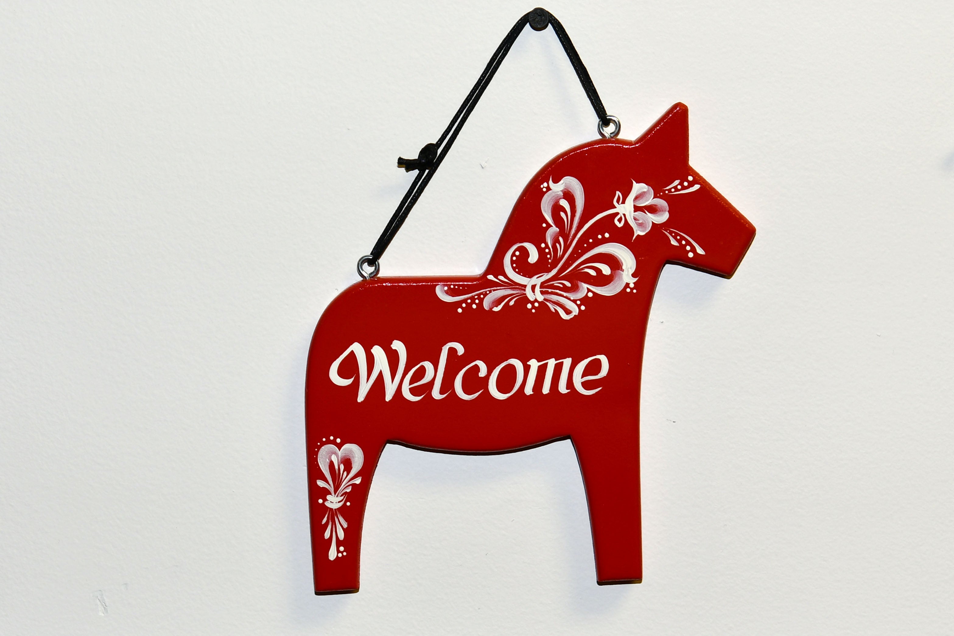 The Sign: Swedish Dala Horse Welcome Wall Decor is a handcrafted, red horse-shaped sign with white patterns and the word Welcome, hanging on a black strap against a white background.