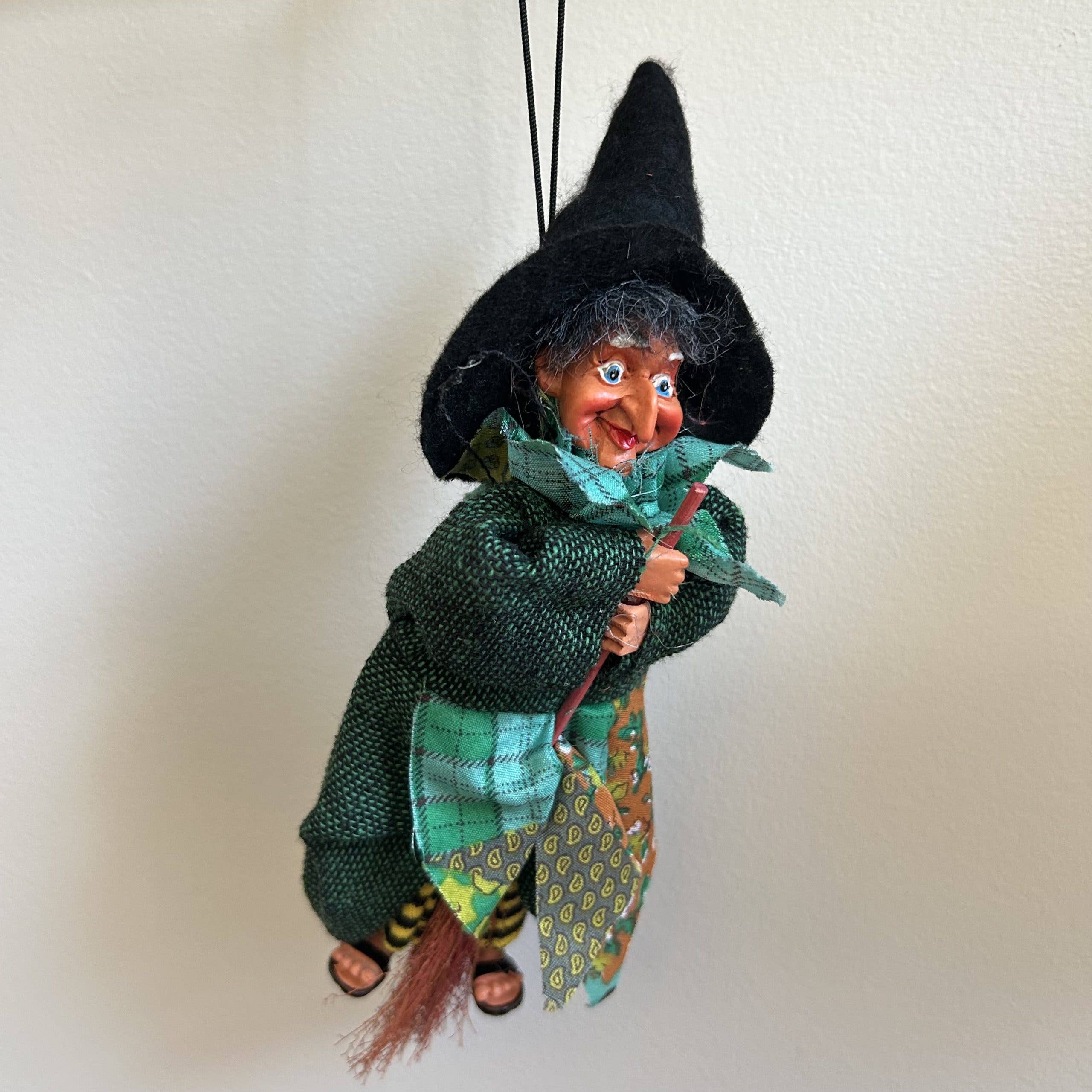 The Kitchen Witch: Johanna Green Kitchen Witch Doll, handmade in Germany, hangs by a black string. She wears a black hat, green outfit, striped leggings, holds a broomstick, and serves as a good luck charm against a white background.