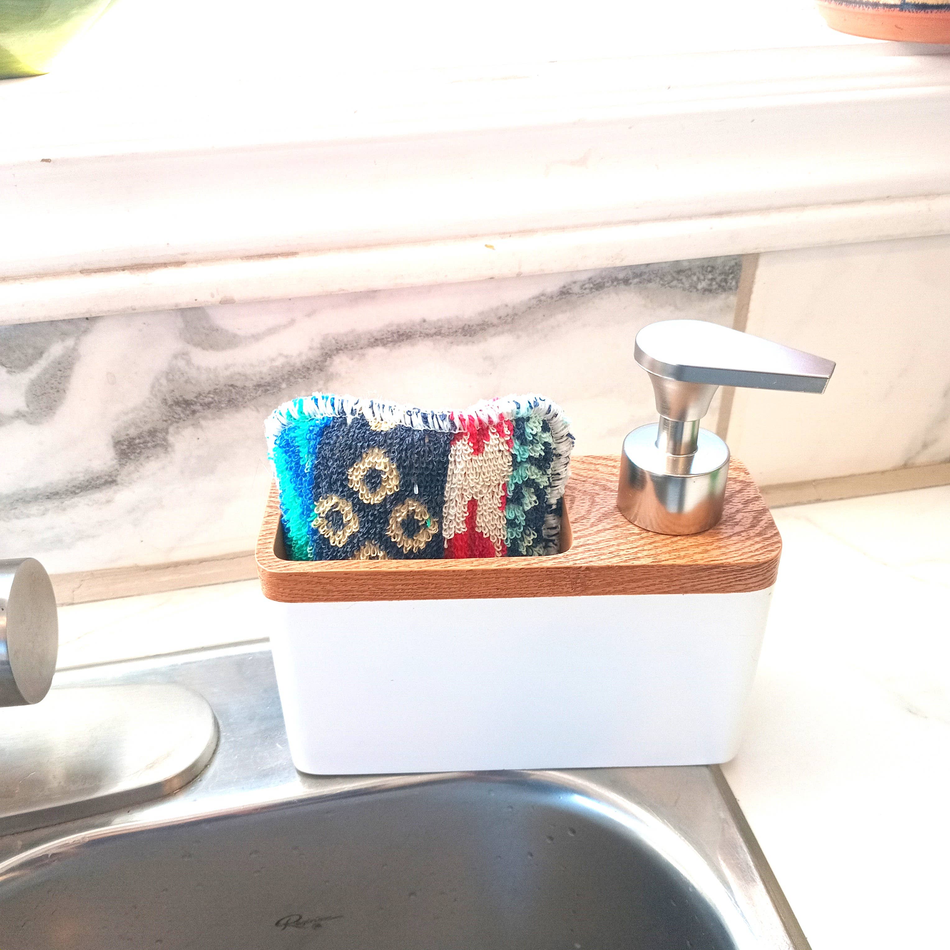 The Scrub and Soap Dispenser combines a soap dispenser with a scrubby holder, organizing your colorful multipurpose scrubby neatly on the kitchen counter next to the sink.