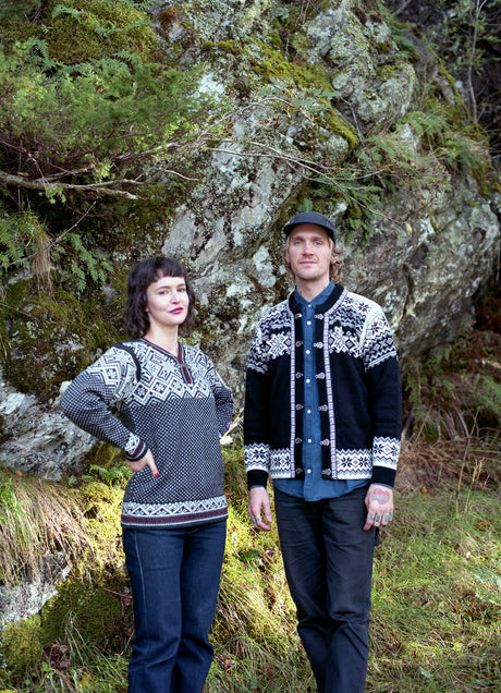A woman and a man stand outdoors in front of a mossy rock and greenery, wearing the Stavanger Norwegian Wool Cardigan in black.